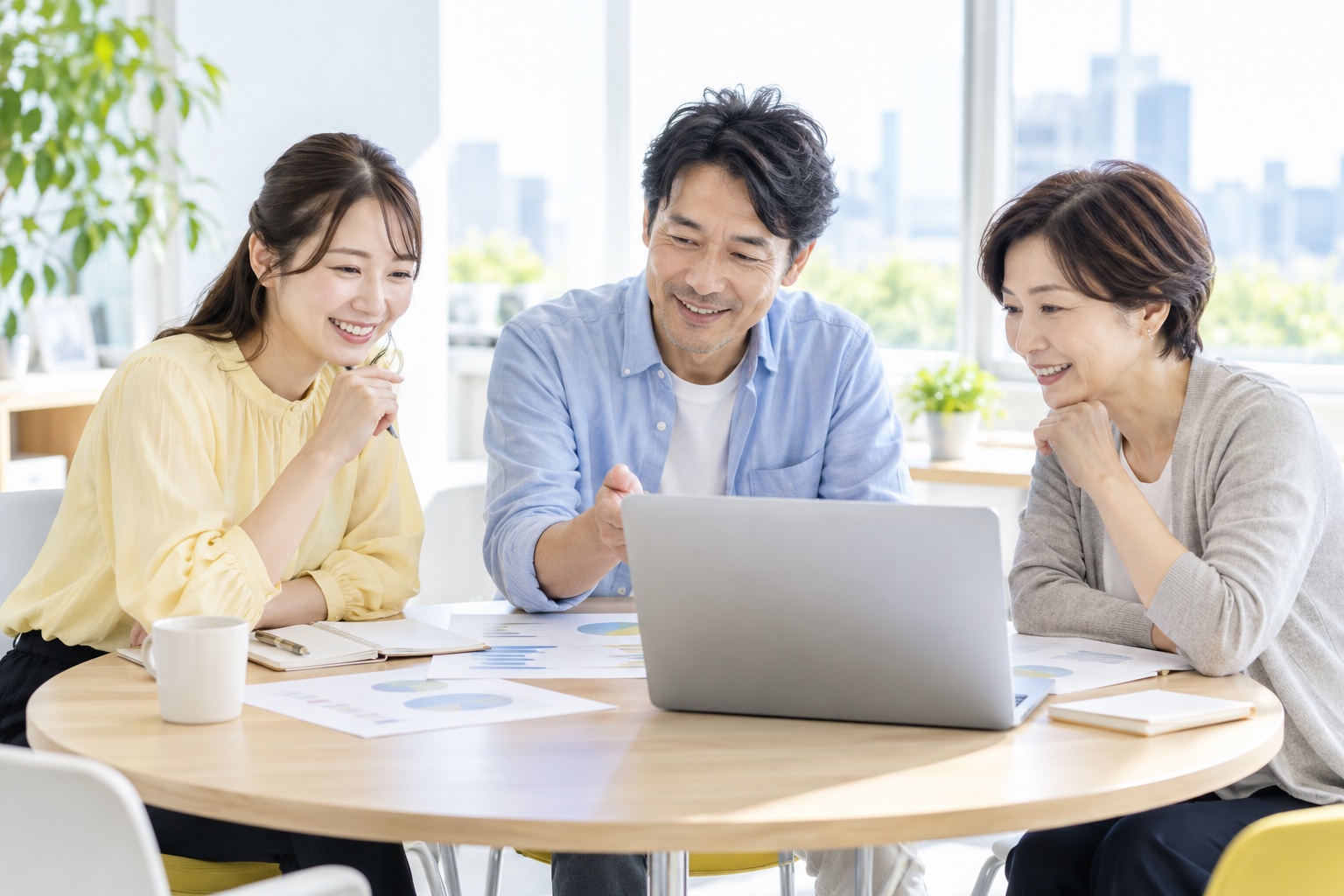 明るいオフィスの丸テーブルで、ノートパソコンを囲んで記事の企画を話し合う年代の違う3人のフリーランス
