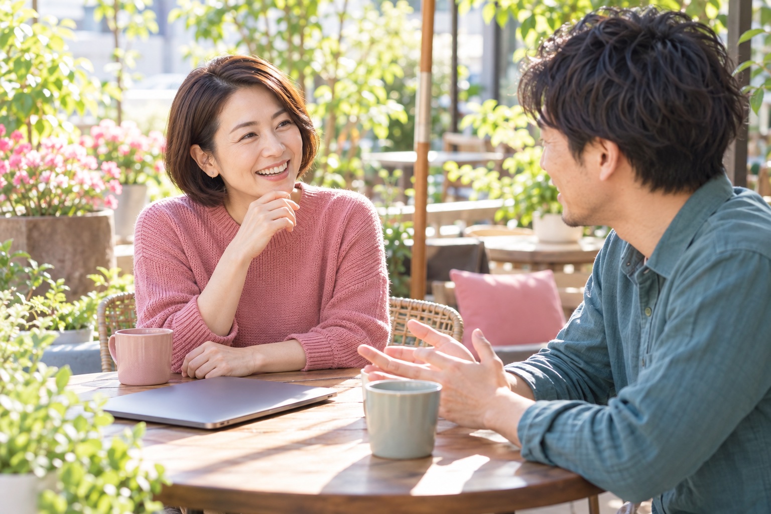 晴れた午前、植物の多いカフェのテラス席で、ノートパソコンを閉じて同業の仲間と笑顔で話す40代の女性ライター