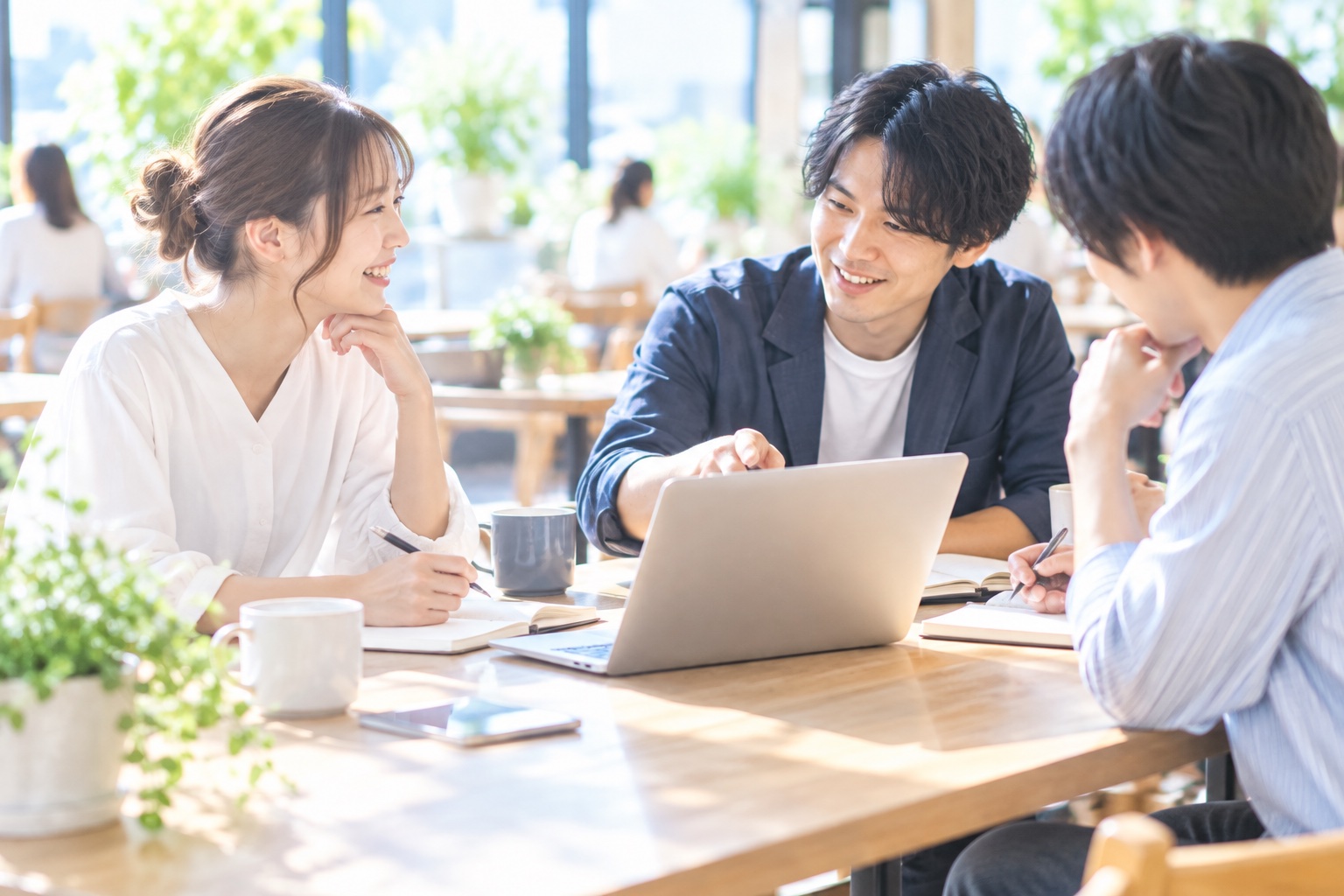 カフェのテーブルでフリーランス仲間3人がノートパソコンを囲み、仕事の分担を話し合っている様子