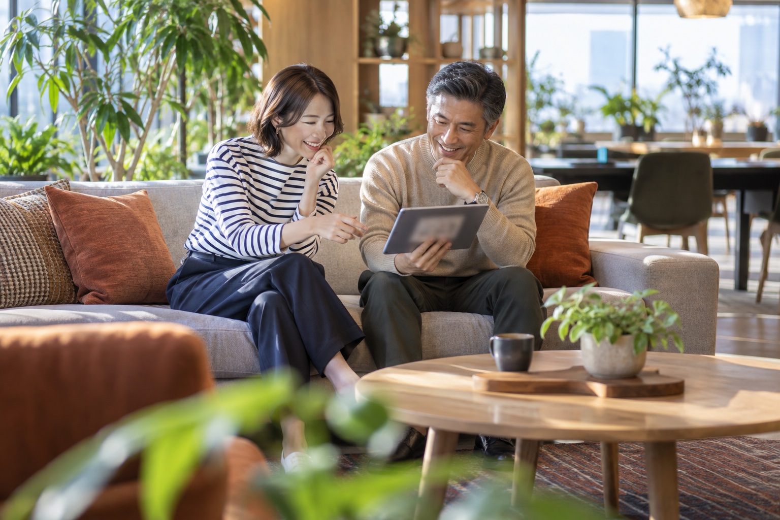 昼下がりのシェアオフィスのラウンジで、ソファに並んで座りタブレットを見ながら笑顔で話す30代女性と50代男性のフリーランス