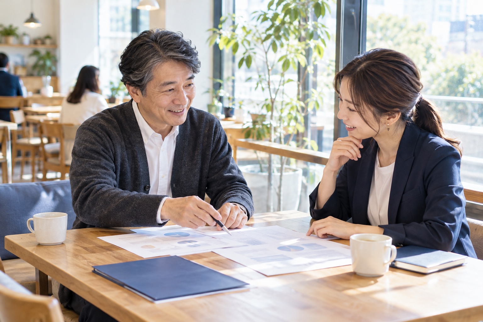 平日の昼、明るいカフェの窓際のテーブルで、取引先の担当者と向かい合って笑顔で打ち合わせをする50代前半の男性フリーランス