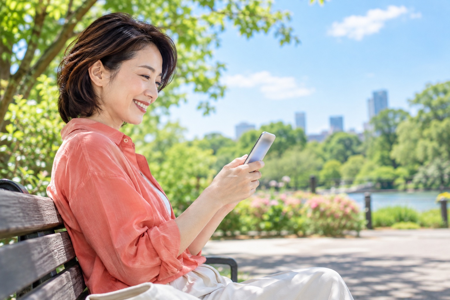 晴れた午前、明るい公園のベンチで、スマホで取引先からのメッセージを読みながら笑顔になる40代の女性フリーランス