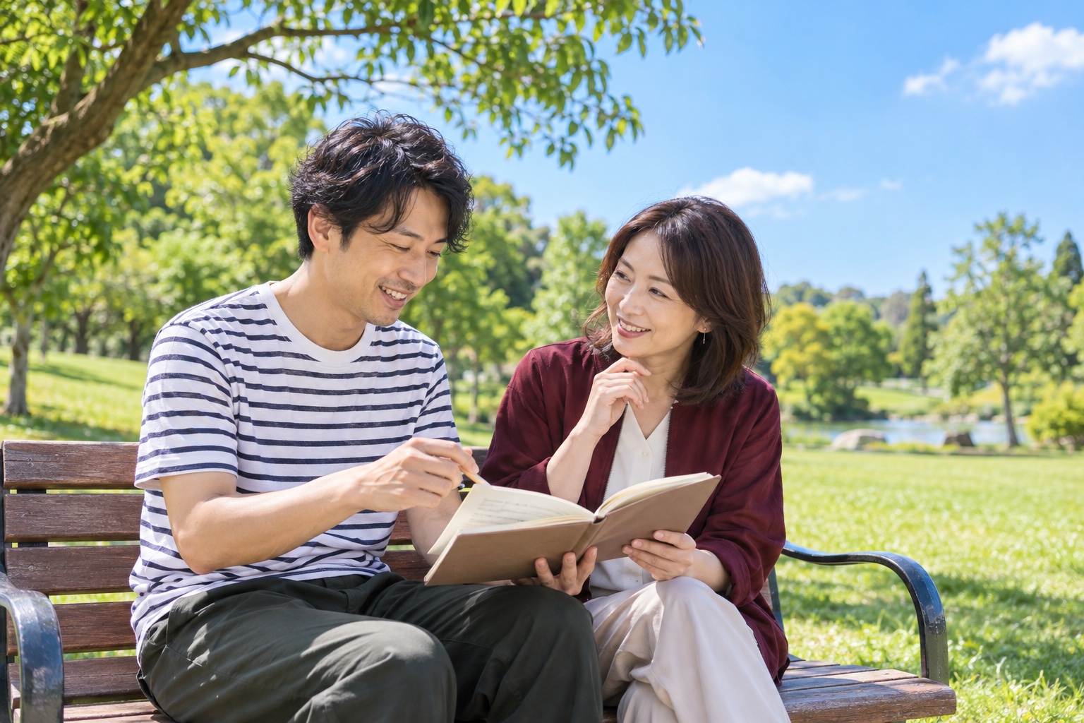 晴れた昼、緑の多い公園のベンチで、1冊のノートを一緒に見ながら仕事の相談をする30代の男性と40代の女性のフリーランス