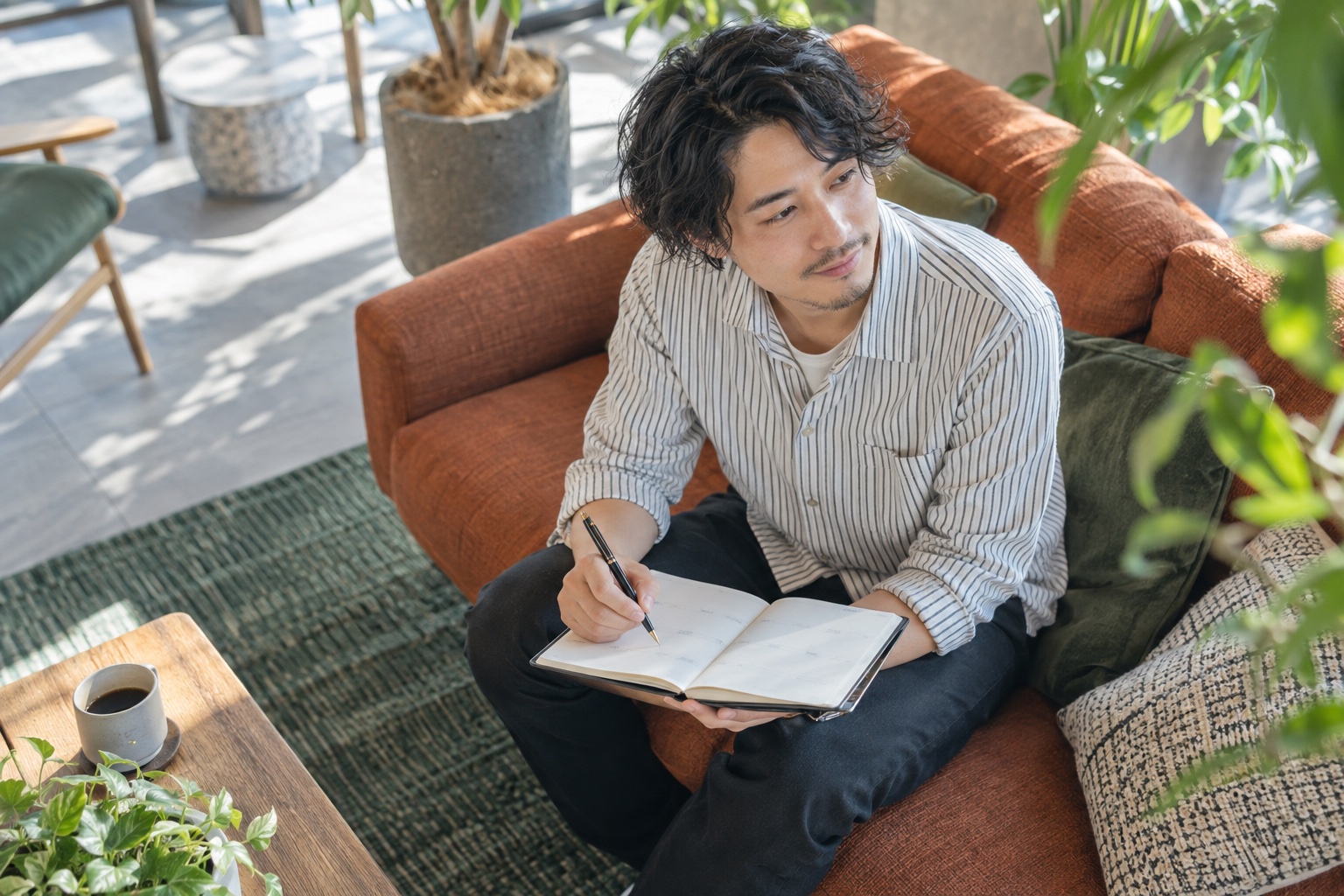 植物に囲まれたコワーキングスペースのソファで、手帳に法人化したい理由を書き出して考える30代の男性フリーランス
