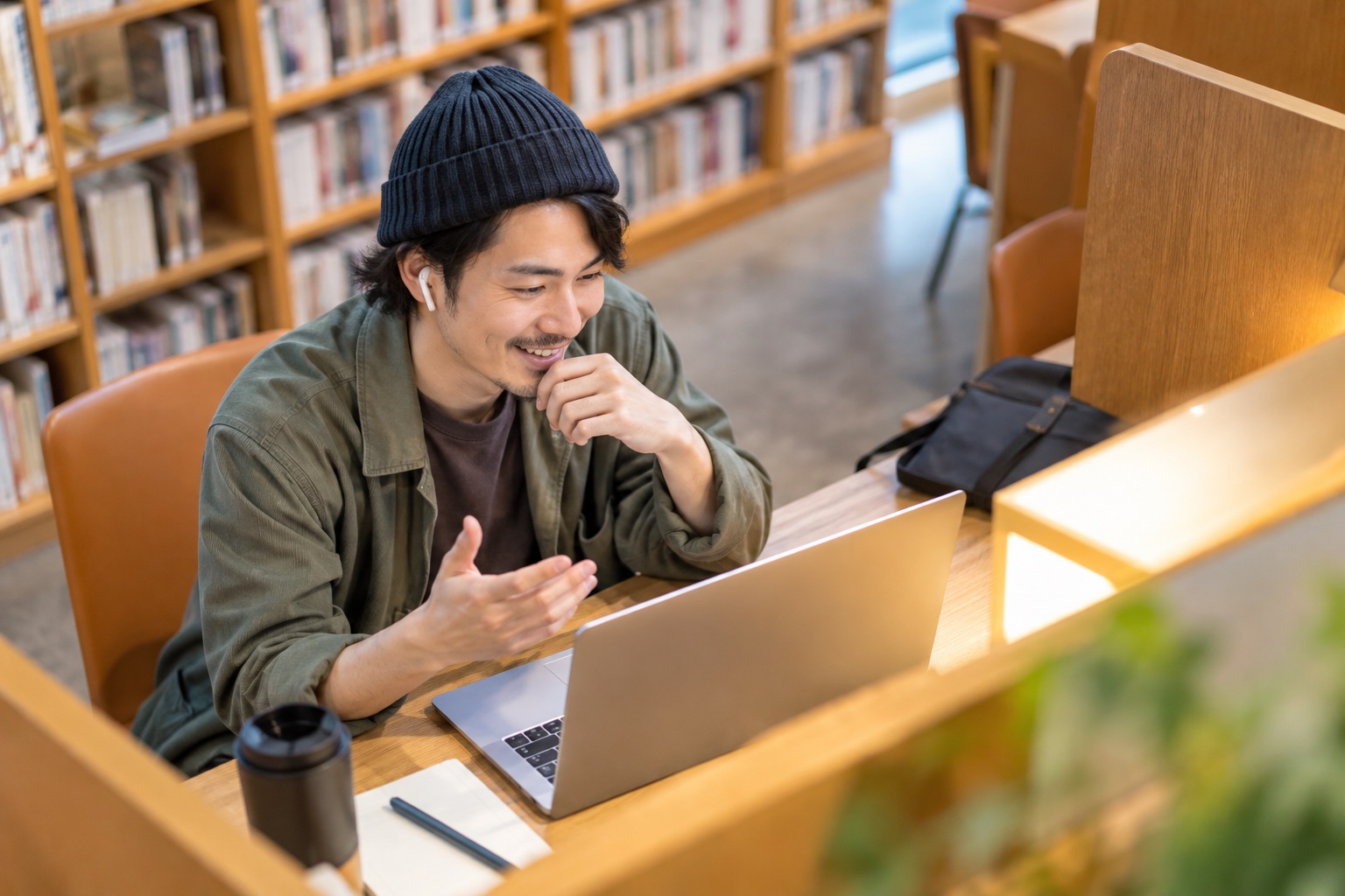図書館の自習ブースで、イヤホンをつけて仲間とオンライン通話で笑顔で話す30代男性のフリーランス