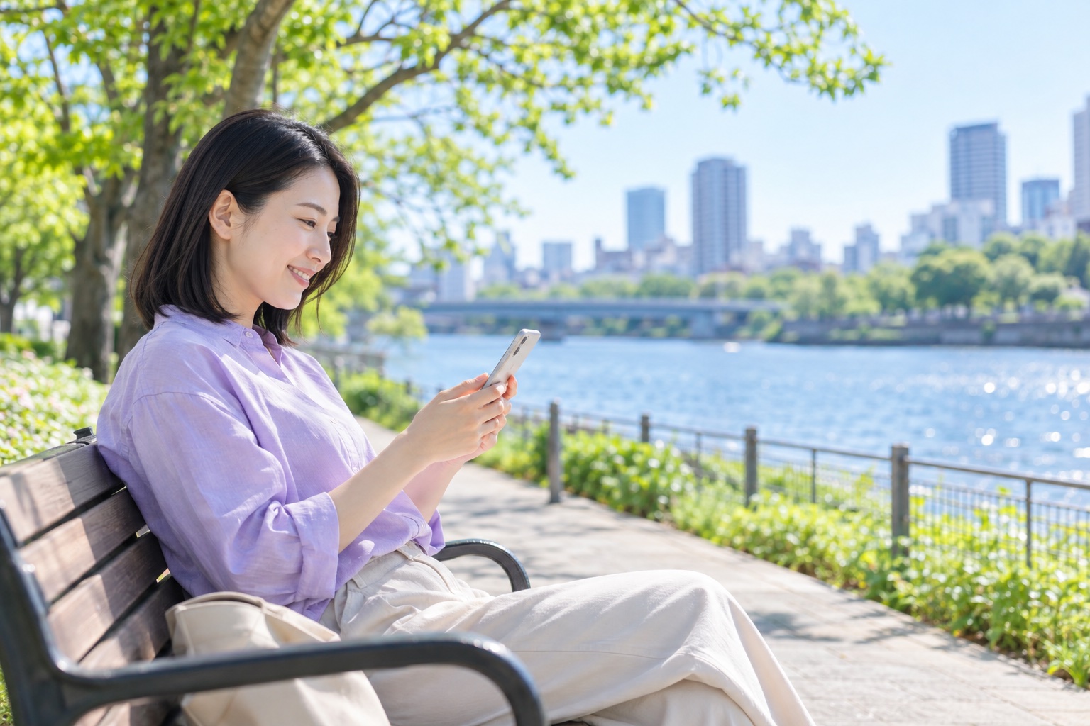 晴れた午前、川沿いの遊歩道のベンチで、仲間からのメッセージをスマホで読んでほほえむ30代の女性フリーランス